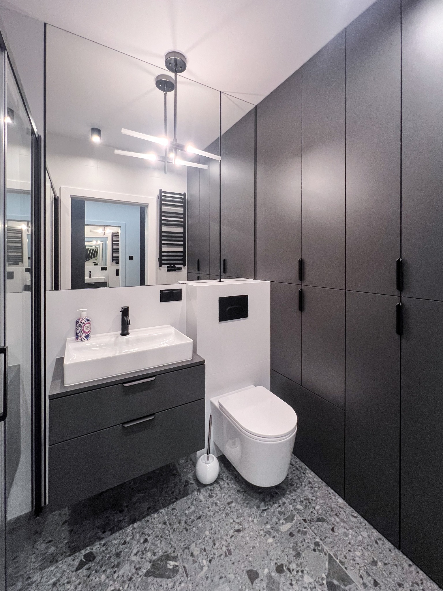 Nowoczesna łazienka z szarą zabudową meblową, umywalką nablatową, toaletą wiszącą i lustrem na całej ścianie, odbijającym wnętrze. Na podłodze terazzo.