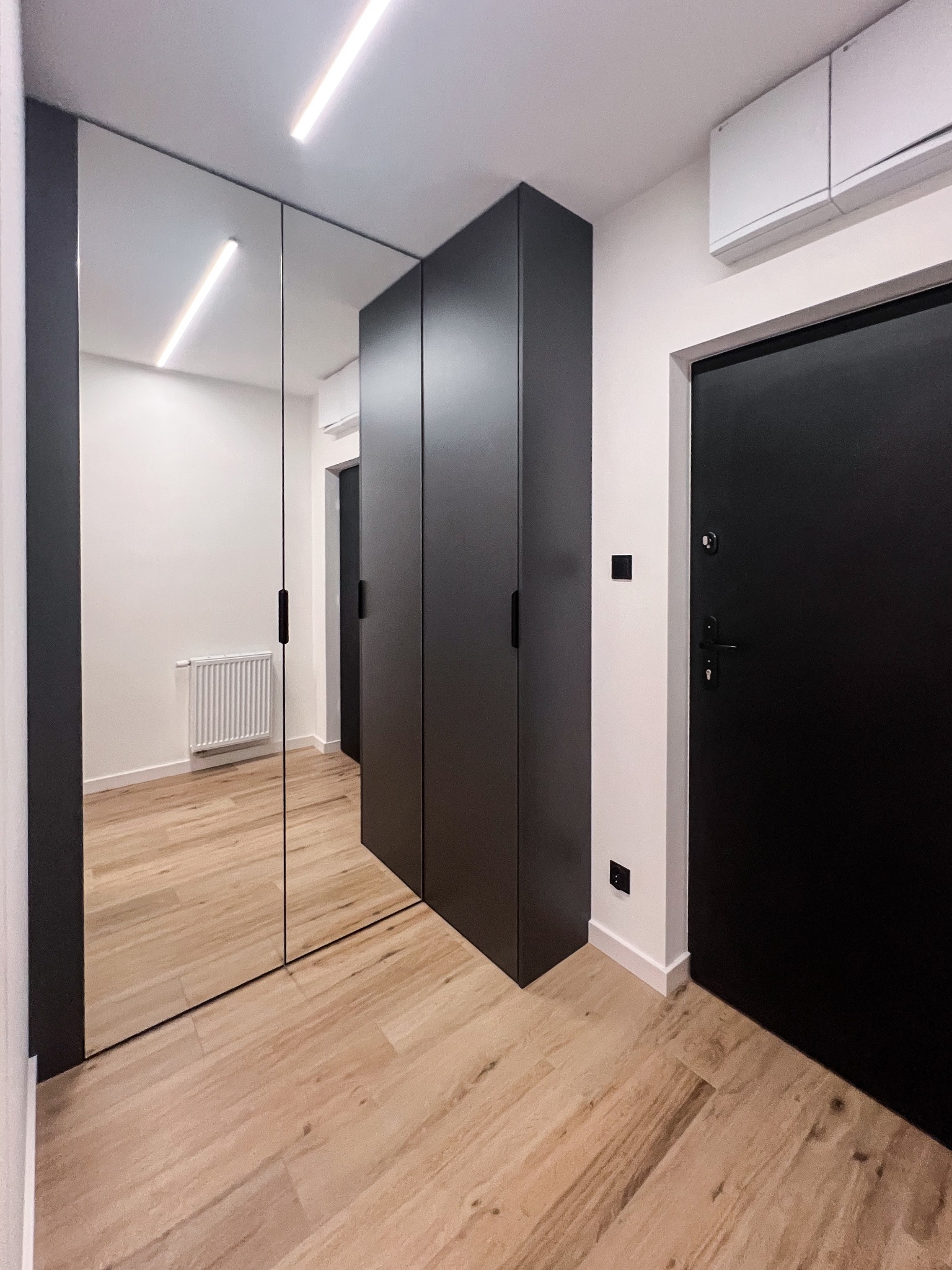 Nowoczesna, ciemnoszara szafa z lustrzanymi drzwiami przesuwnymi w przedpokoju z drewnopodobną podłogą i czarnymi drzwiami wejściowymi, oświetlenie LED na suficie.