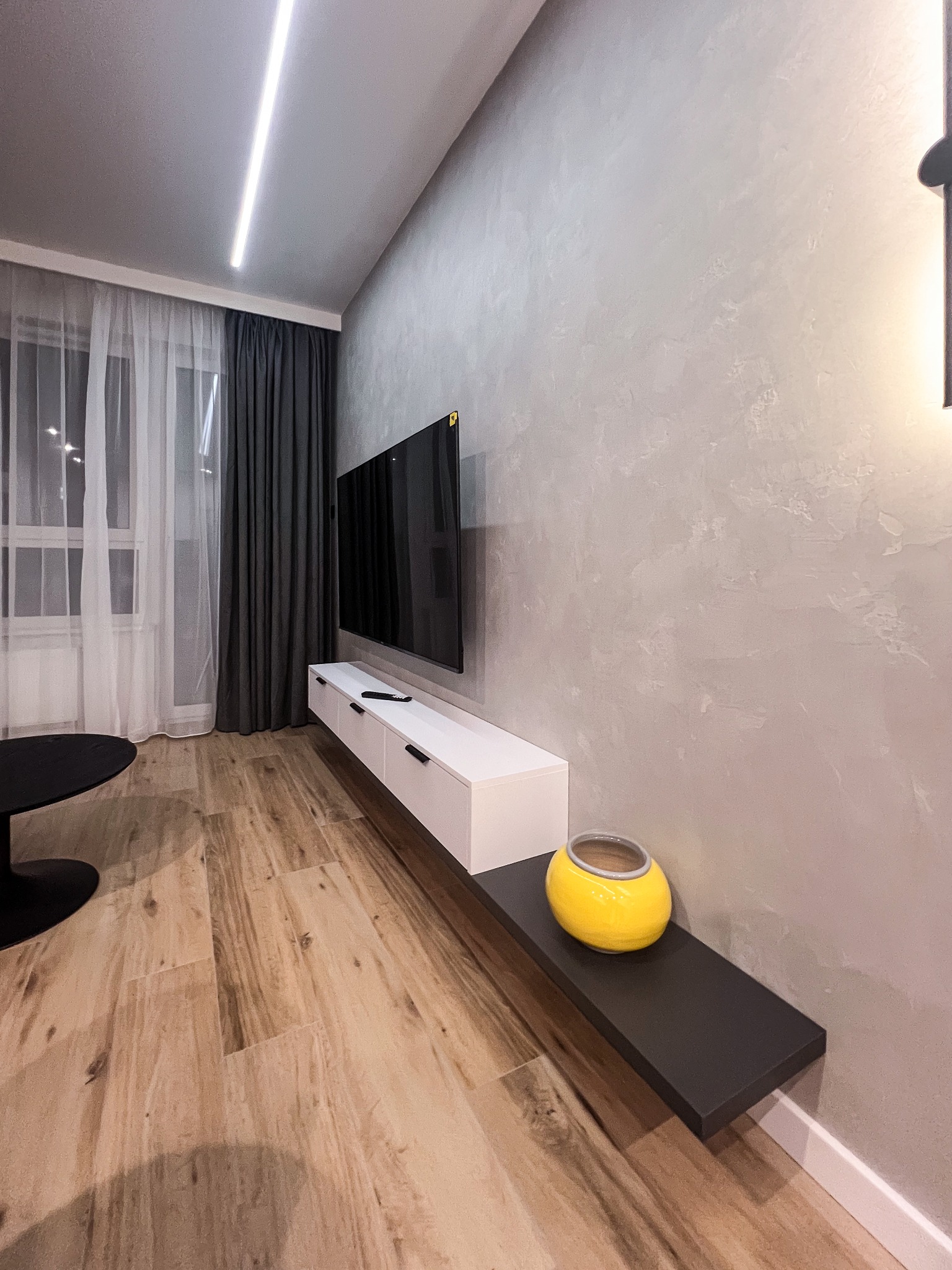 Nowoczesny salon z podwieszaną białą szafką RTV, telewizorem na ścianie, drewnianą podłogą i szarą ścianą z dekoracyjnym tynkiem.