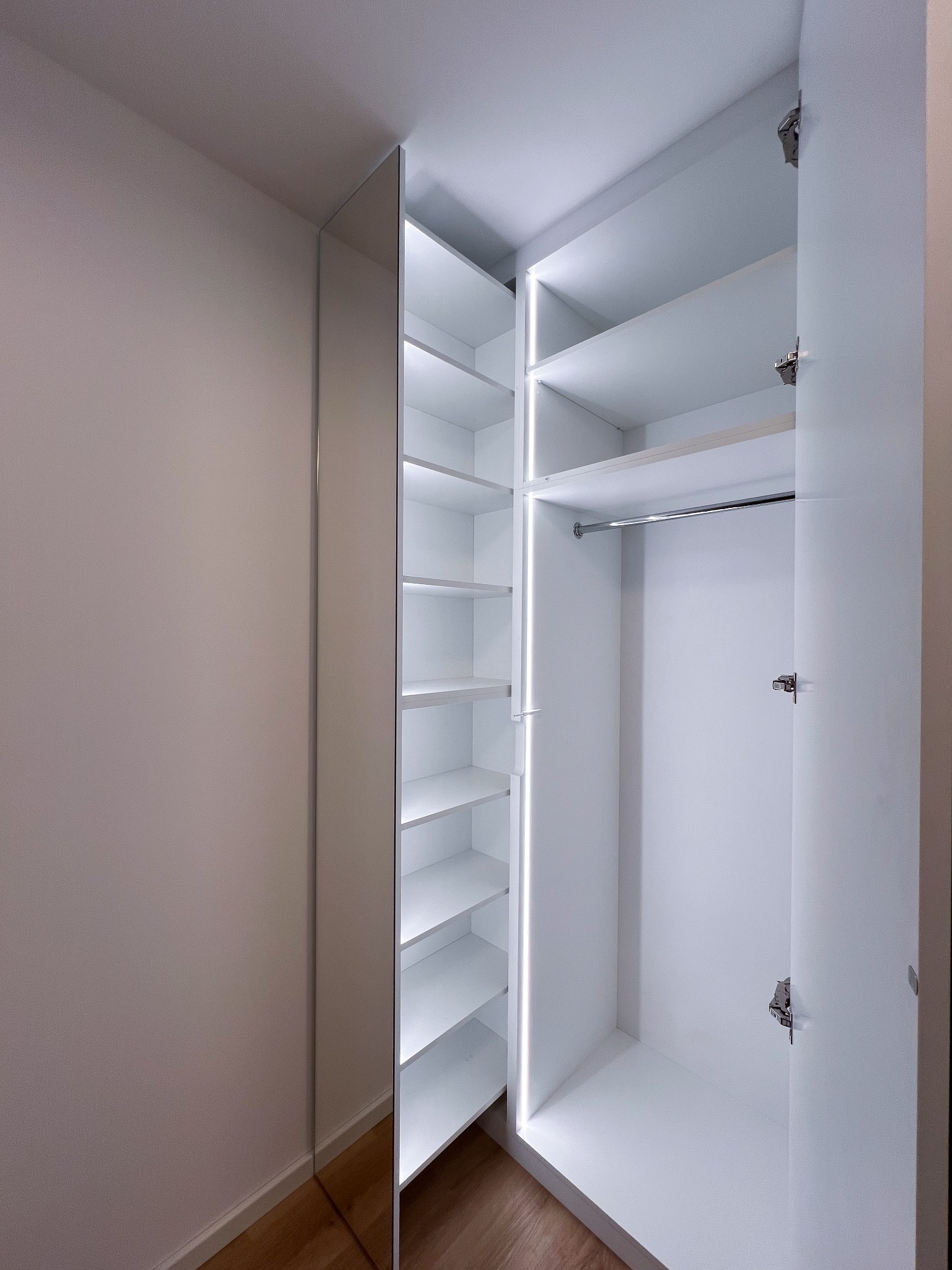 Wnętrze otwartej, białej szafy wnękowej z oświetleniem LED we Wrocławiu, widoczny system półek i drążek na ubrania, z lustrzanym frontem po lewej stronie.