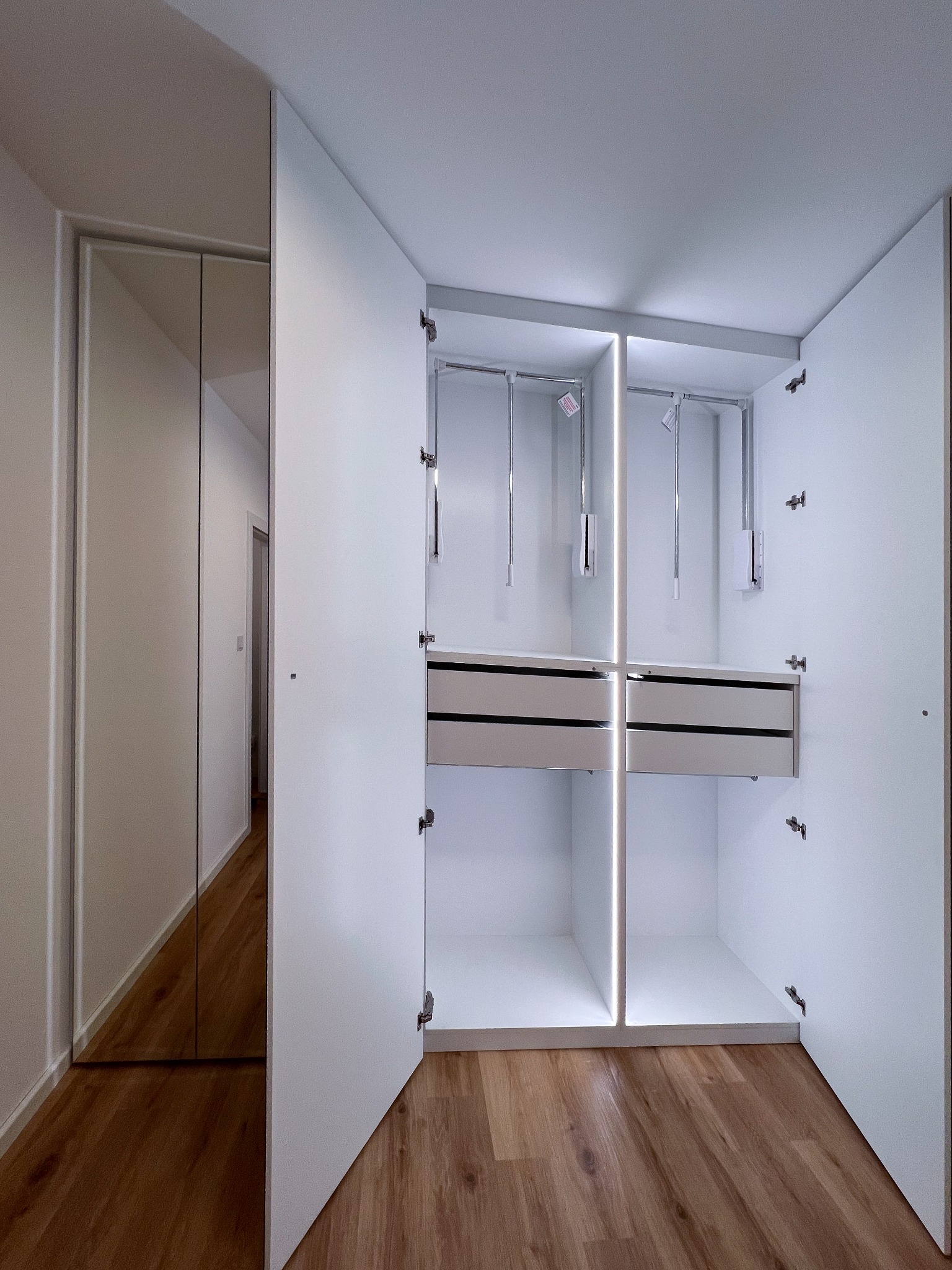 Wnętrze białej, otwartej szafy z szufladami i drążkami na ubrania, podświetlane pionowymi listwami LED, obok szafa z lustrzanymi drzwiami.