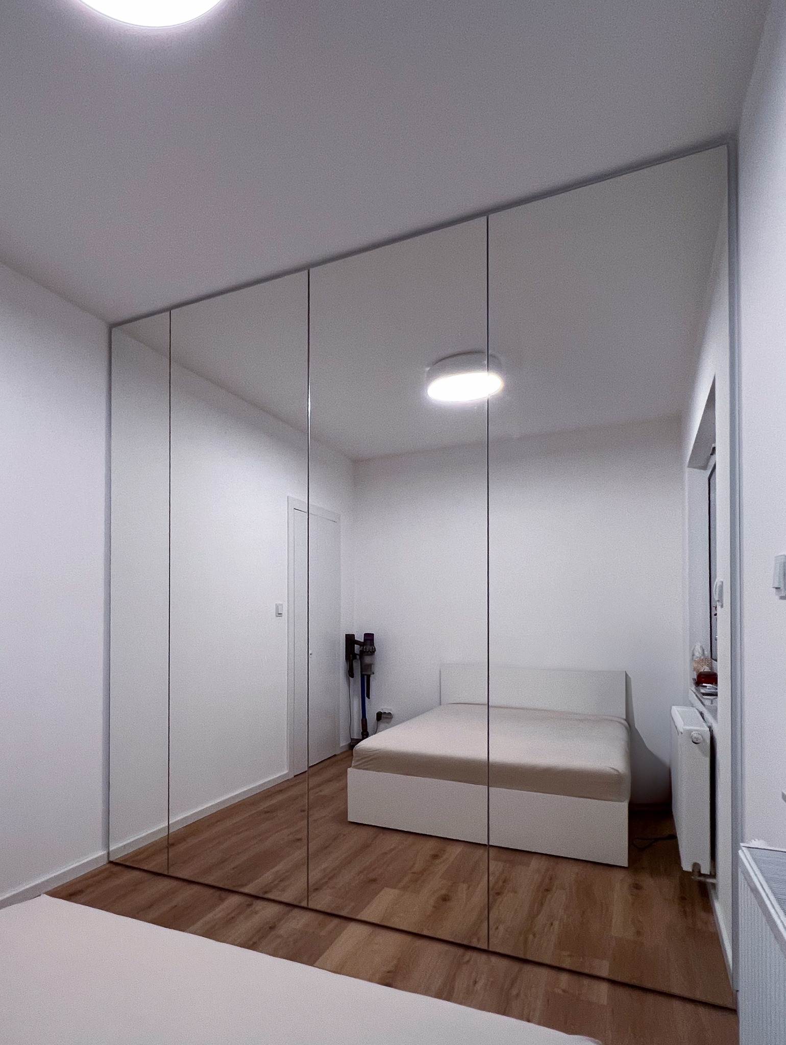 Nowoczesna szafa wnękowa z lustrzanymi drzwiami przesuwnymi, odbijająca wnętrze sypialni z łóżkiem i drewnianą podłogą.