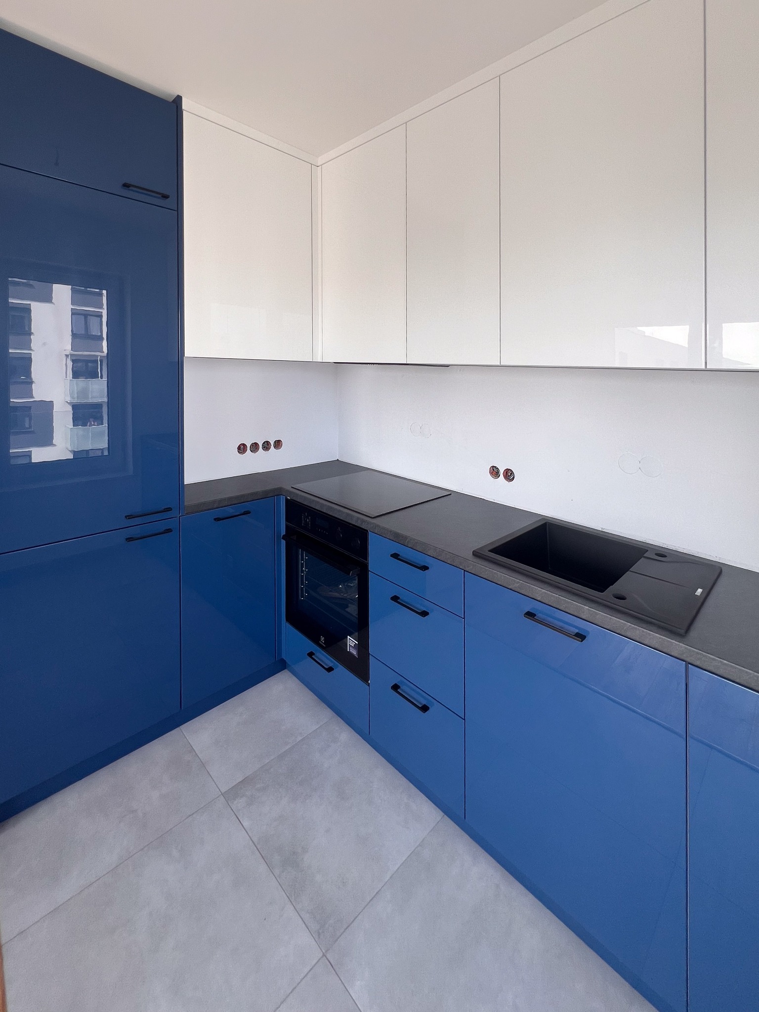 Nowoczesna kuchnia z niebieskimi, lakierowanymi szafkami dolnymi i białymi górnymi, szary blat, czarny zlew i płyta grzewcza, widoczne niedokończone gniazdka elektryczne na białej ścianie.