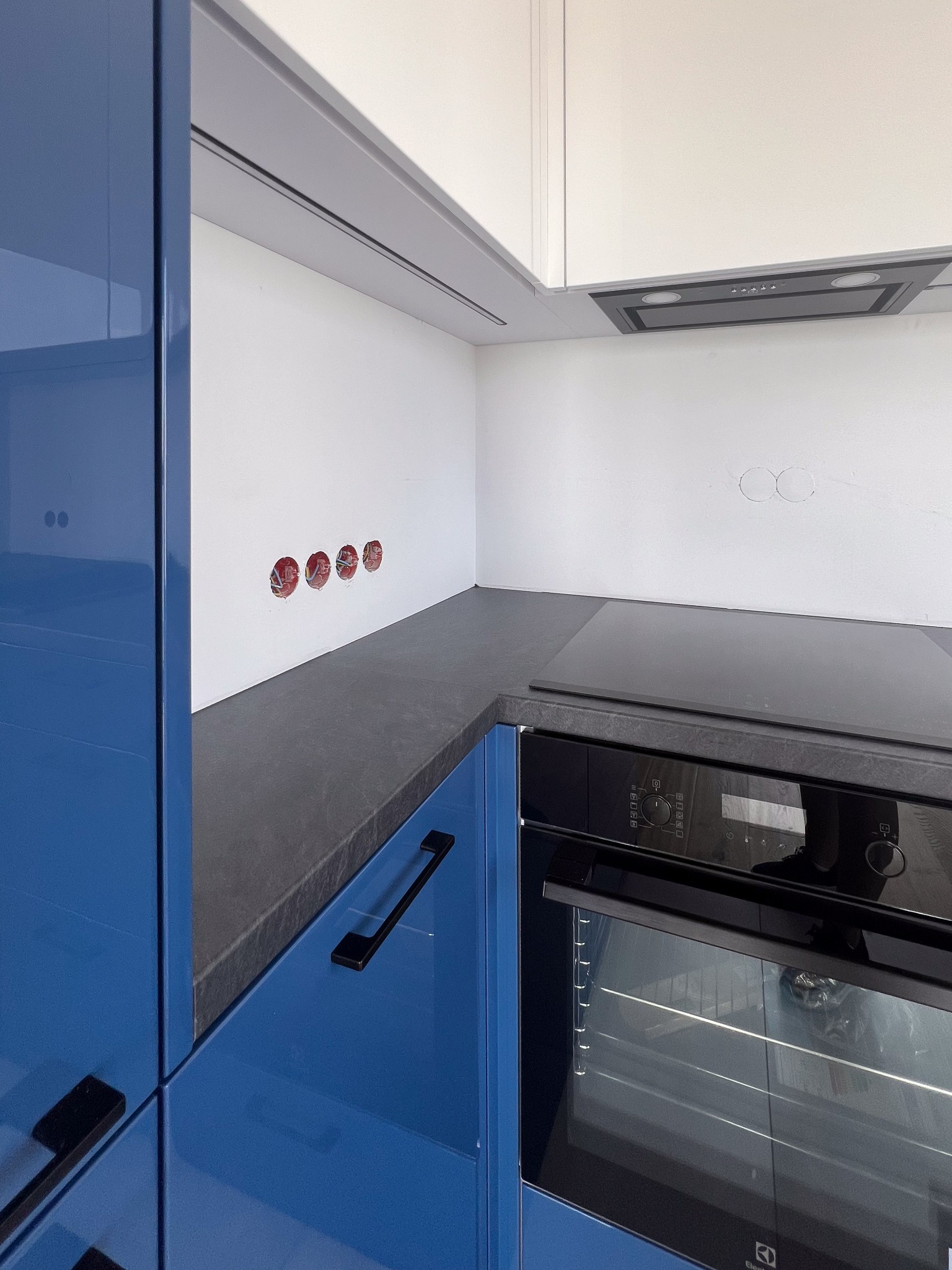 Fragment kuchni: niebieskie, lakierowane szafki dolne z czarnymi uchwytami, szary blat, białe szafki górne, płyta indukcyjna i piekarnik Electrolux. Widoczne gniazdka elektryczne w ścianie.