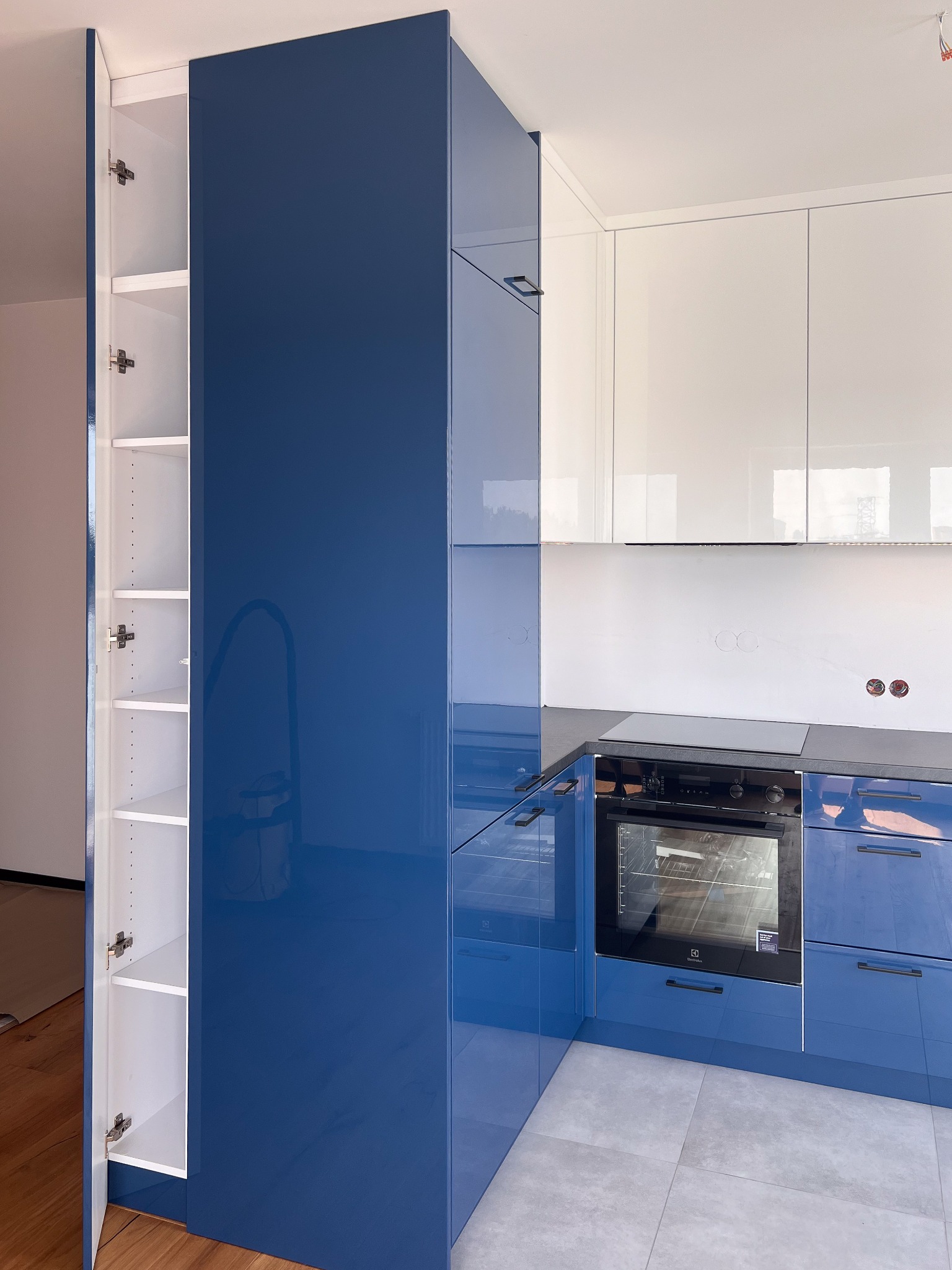 Nowoczesna kuchnia z błyszczącymi, granatowymi szafkami dolnymi i białymi szafkami górnymi, otwarty słupek z półkami po lewej stronie, płyta indukcyjna i piekarnik Electrolux w zabudowie.
