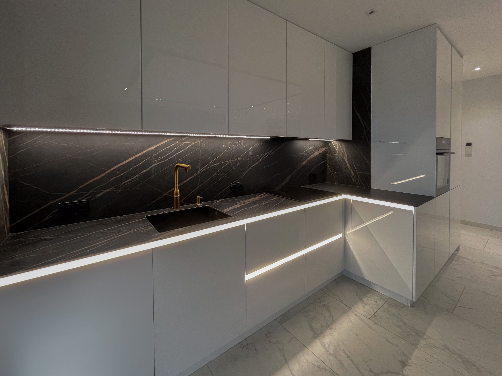 Nowoczesna kuchnia z białymi, lakierowanymi frontami, czarnym blatem imitującym marmur i złotą armaturą, oświetlona listwami LED pod szafkami wiszącymi i między frontami szafek dolnych.