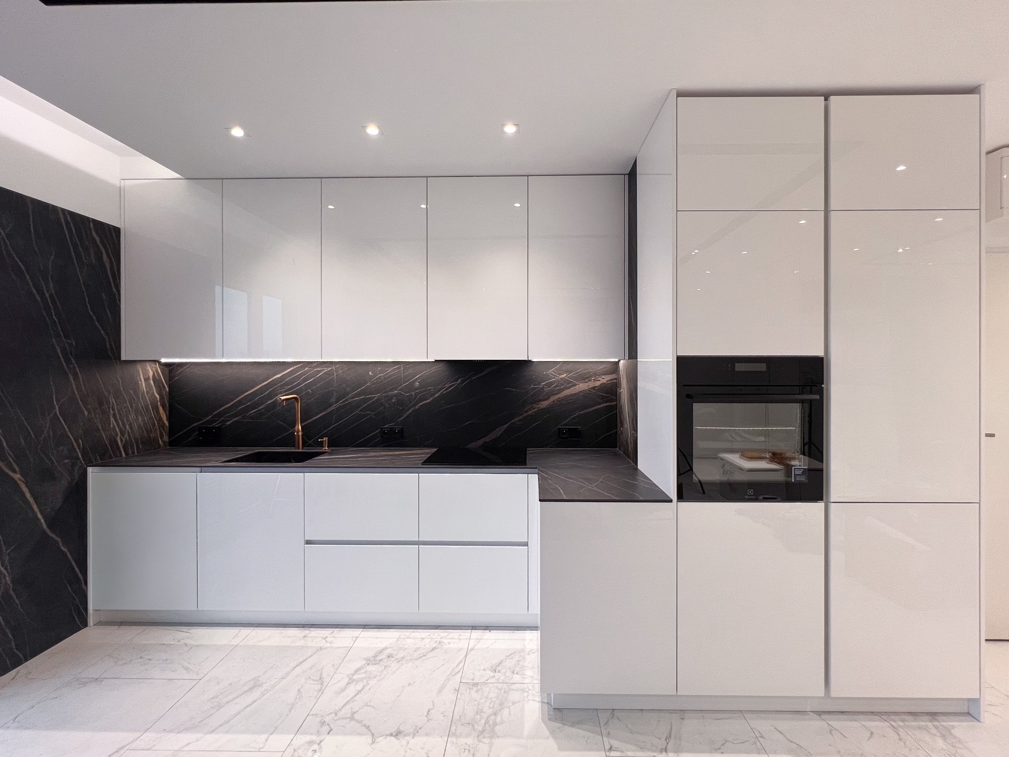 Nowoczesna kuchnia z białymi, lakierowanymi frontami, czarnym blatem z marmurowym wzorem i złotą armaturą, oświetleniem LED pod szafkami i marmurową podłogą.