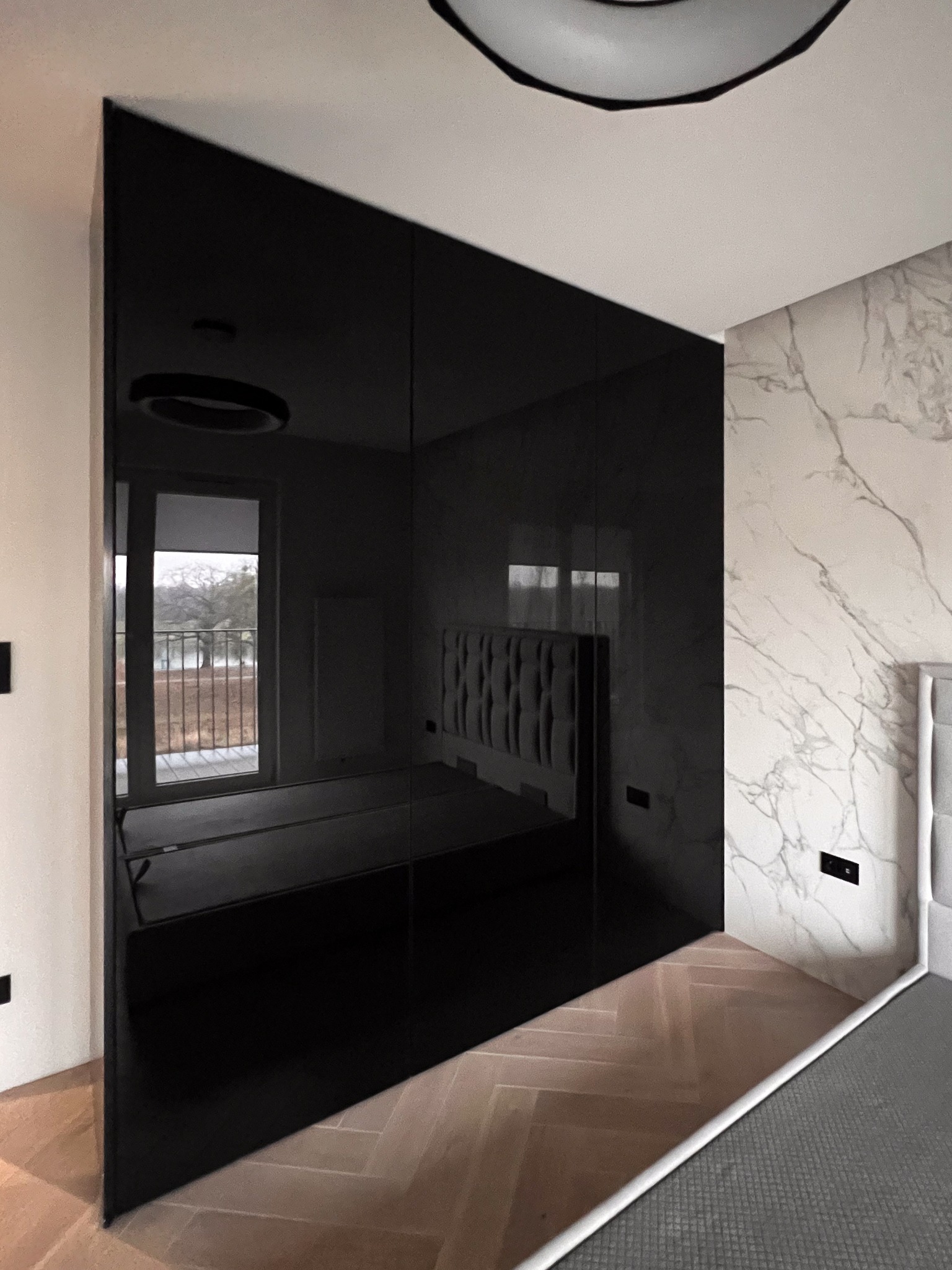 Czarna, błyszcząca zabudowa wnękowa z lustrzanymi drzwiami przesuwnymi, odbijająca wnętrze sypialni z łóżkiem i balkonem, na tle ściany z marmurowym wzorem i parkietem w jodełkę.