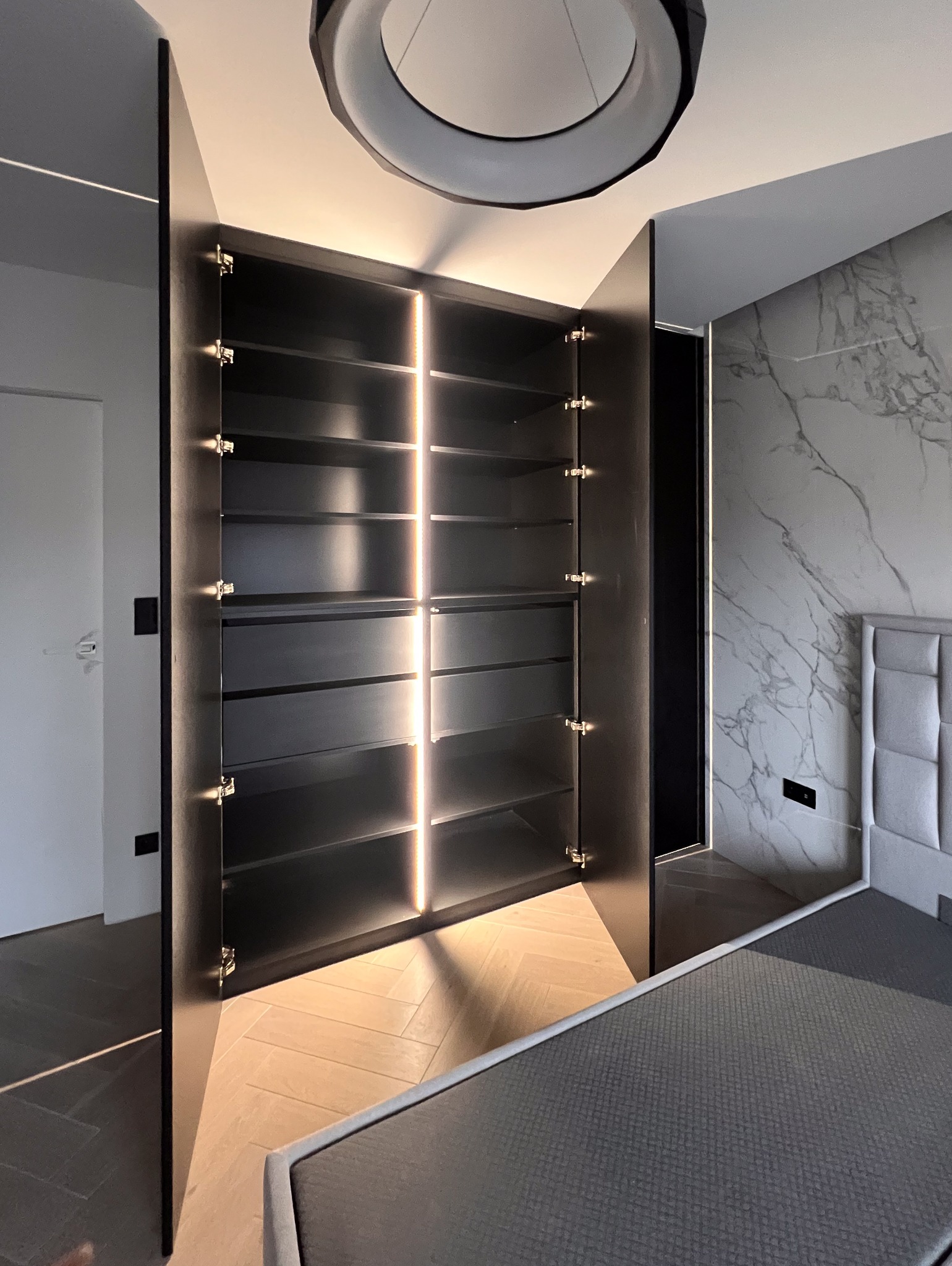 Otwarta, czarna szafa z podświetlanymi półkami w nowoczesnej sypialni z marmurową ścianą i parkietem w jodełkę.