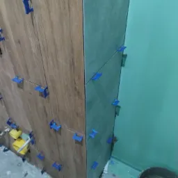 Proces kładzenia płytek imitujących drewno na ścianie, z użyciem niebieskich klipsów poziomujących, widoczny fragment ściany pomalowanej na zielono oraz wiadro z zaprawą.