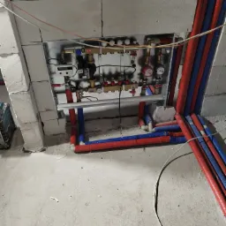 Rozdzielacz ogrzewania podłogowego zamontowany na ścianie z gazobetonu, z widocznymi rurami doprowadzającymi i odprowadzającymi wodę w izolacji czerwonej i niebieskiej, w surowym stanie wykończenia.