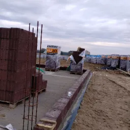 Budowa ogrodzenia z brązowych bloczków betonowych, widoczne zbrojenie narożnika, palety z zapakowanymi bloczkami w tle, kubek na murku.