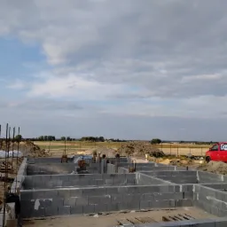 Fundamenty domu z bloczków betonowych na placu budowy, widoczny czerwony samochód dostawczy w tle.