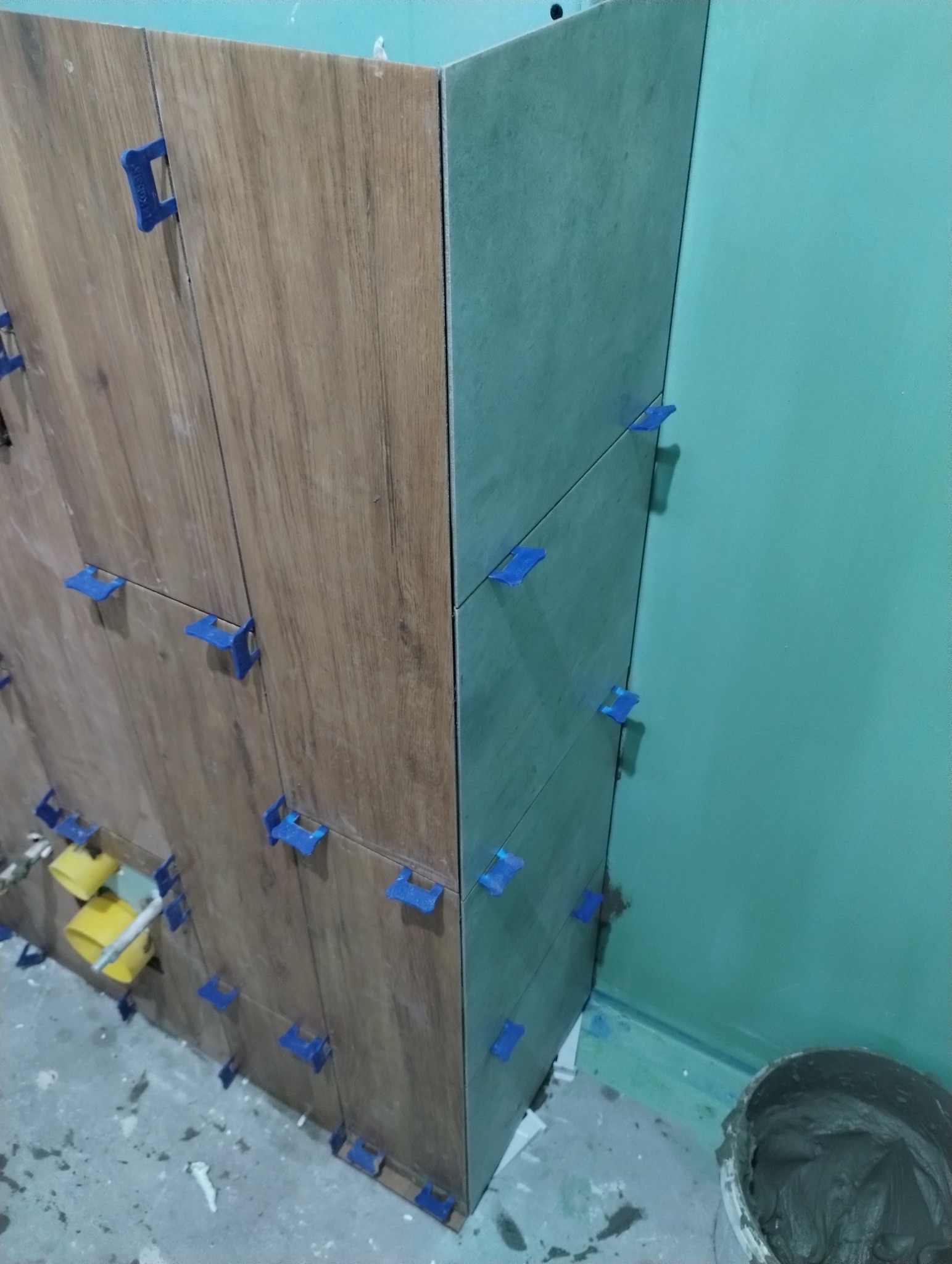 Proces kładzenia płytek imitujących drewno na ścianie, z użyciem niebieskich klipsów poziomujących, widoczny fragment ściany pomalowanej na zielono oraz wiadro z zaprawą.