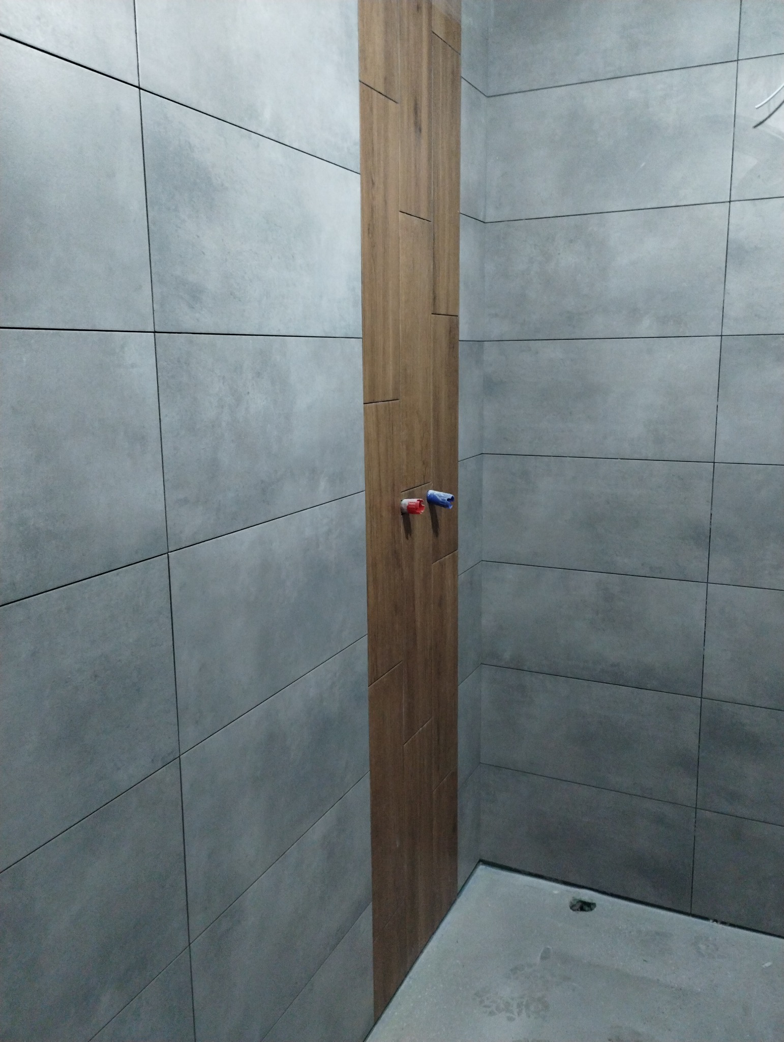 Szara, matowa, prostokątna okładzina ścienna z drewnopodobnym akcentem pionowym i wystającymi przyłączami wodnymi w narożniku łazienki.