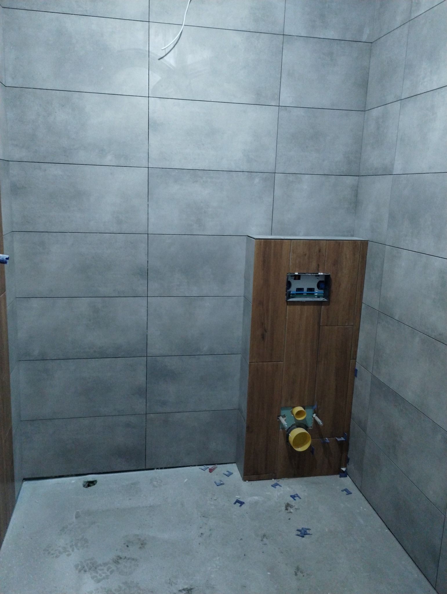 Świeżo wykafelkowana łazienka z szarymi płytkami i zabudową WC w kolorze drewna, widoczne żółte rury instalacyjne i plastikowe krzyżyki dystansowe na podłodze.