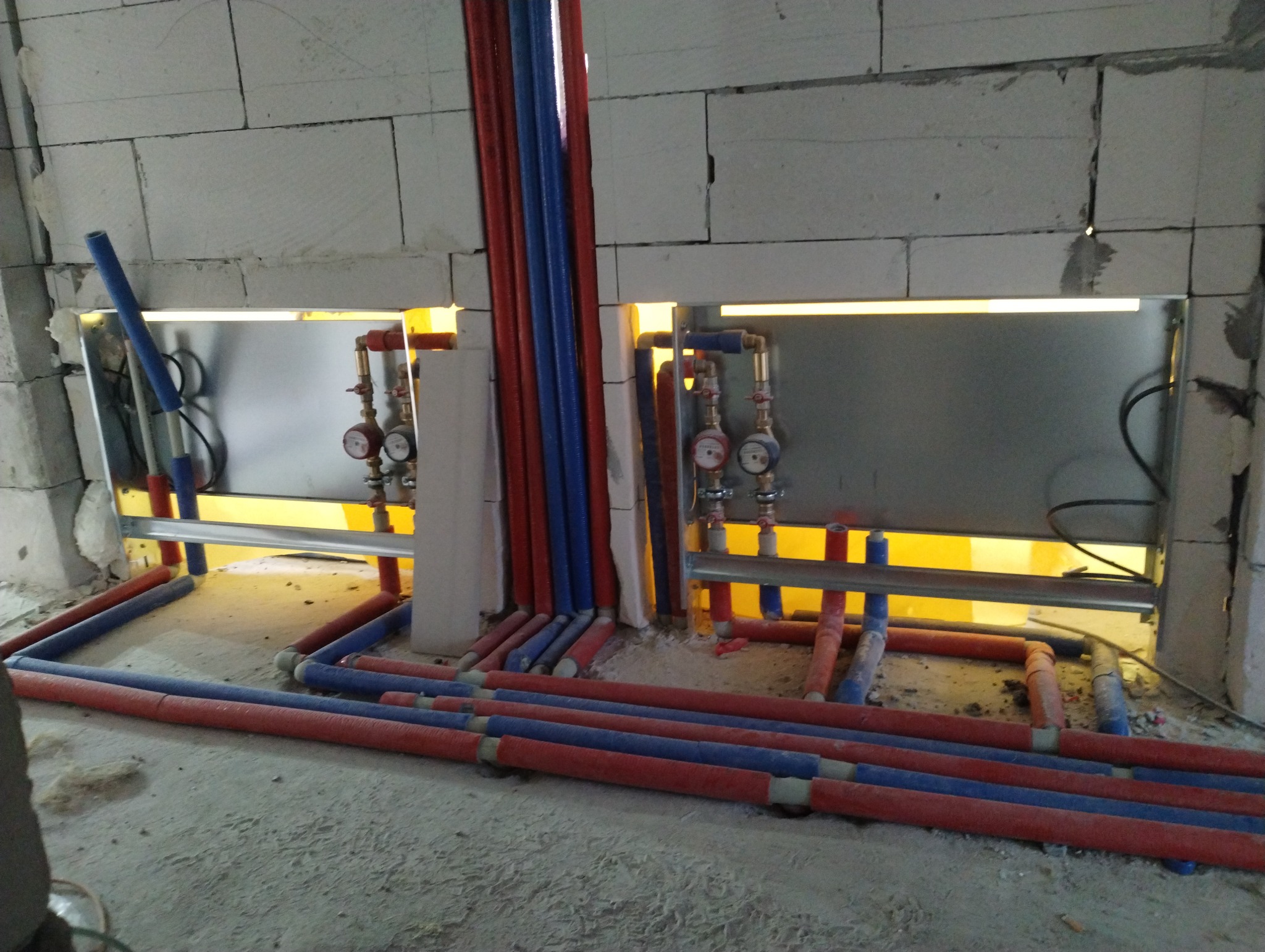 Instalacja wodna w trakcie montażu w surowym stanie, widoczne rury czerwone i niebieskie ułożone na podłodze oraz zawory z manometrami zamontowane w ścianie z podświetleniem.