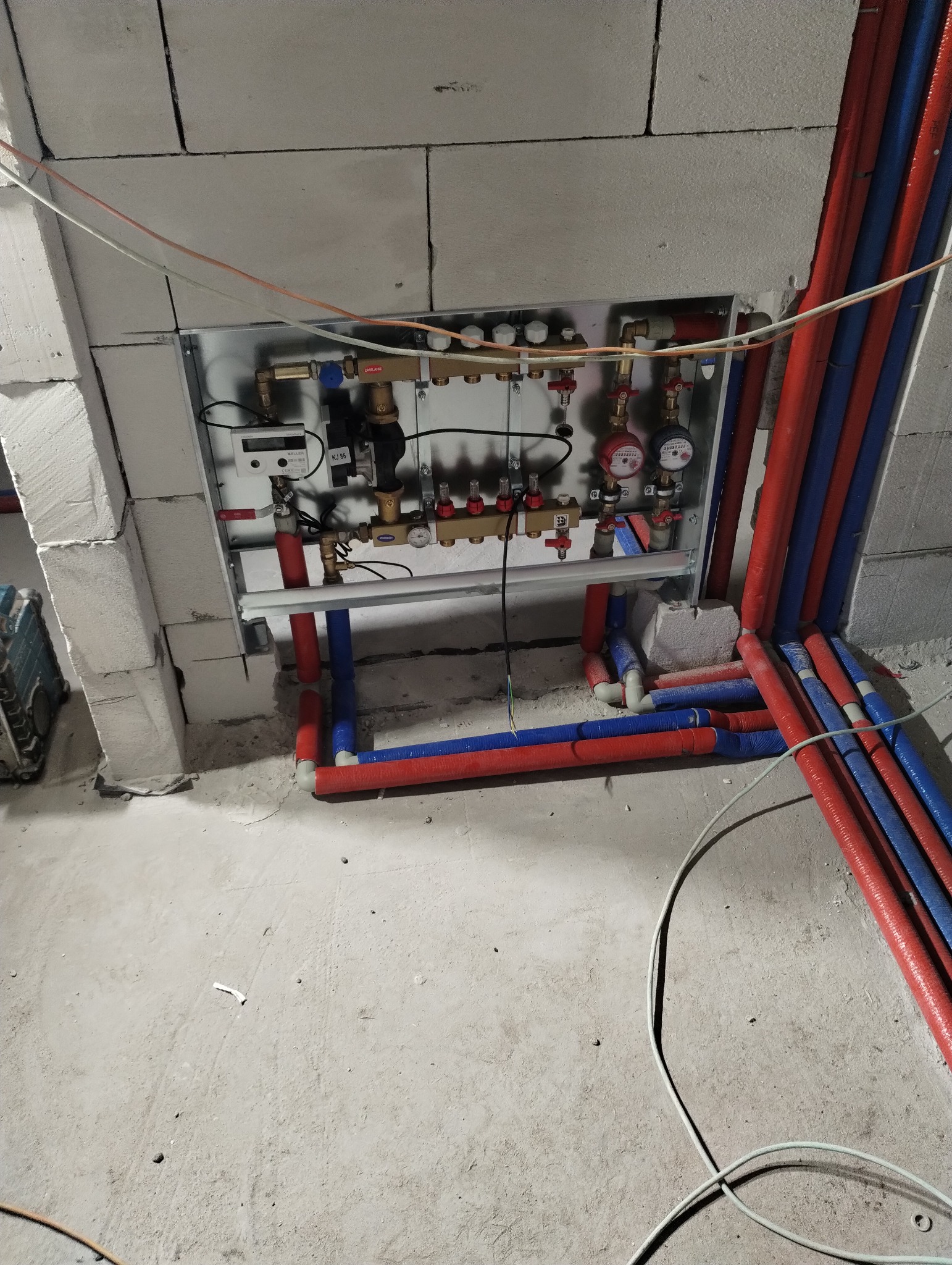 Rozdzielacz ogrzewania podłogowego zamontowany na ścianie z gazobetonu, z widocznymi rurami doprowadzającymi i odprowadzającymi wodę w izolacji czerwonej i niebieskiej, w surowym stanie wykończenia.