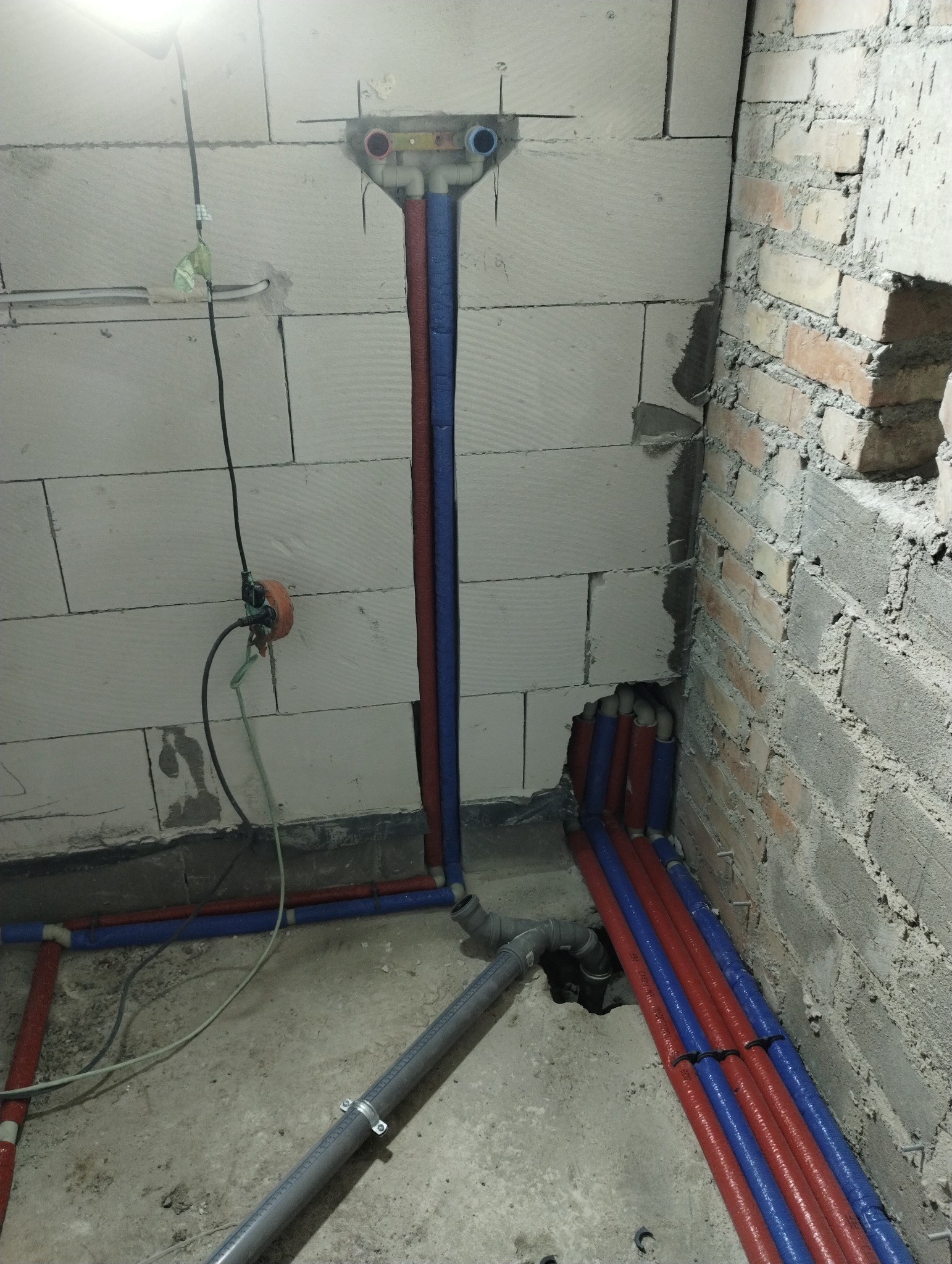 Instalacja wodna w trakcie realizacji w surowym wnętrzu, widoczne rury w izolacji czerwonej i niebieskiej, oraz szara rura kanalizacyjna, ściany z pustaków i cegły.
