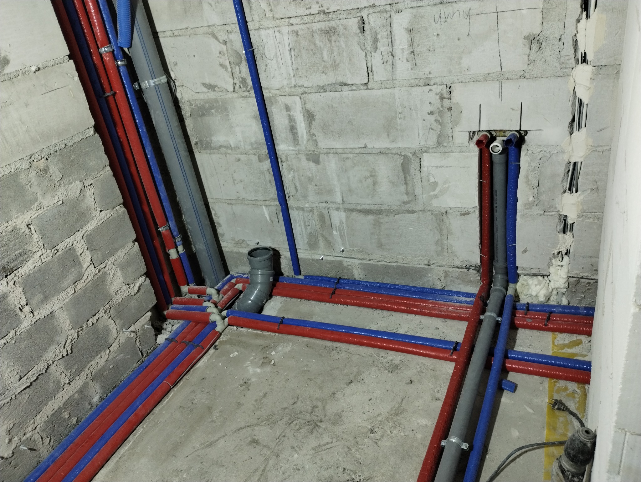 Instalacja wodna i kanalizacyjna w trakcie montażu w surowym wnętrzu łazienki z widocznymi rurami w kolorze czerwonym, niebieskim i szarym, przymocowanymi do ścian z betonu komórkowego.