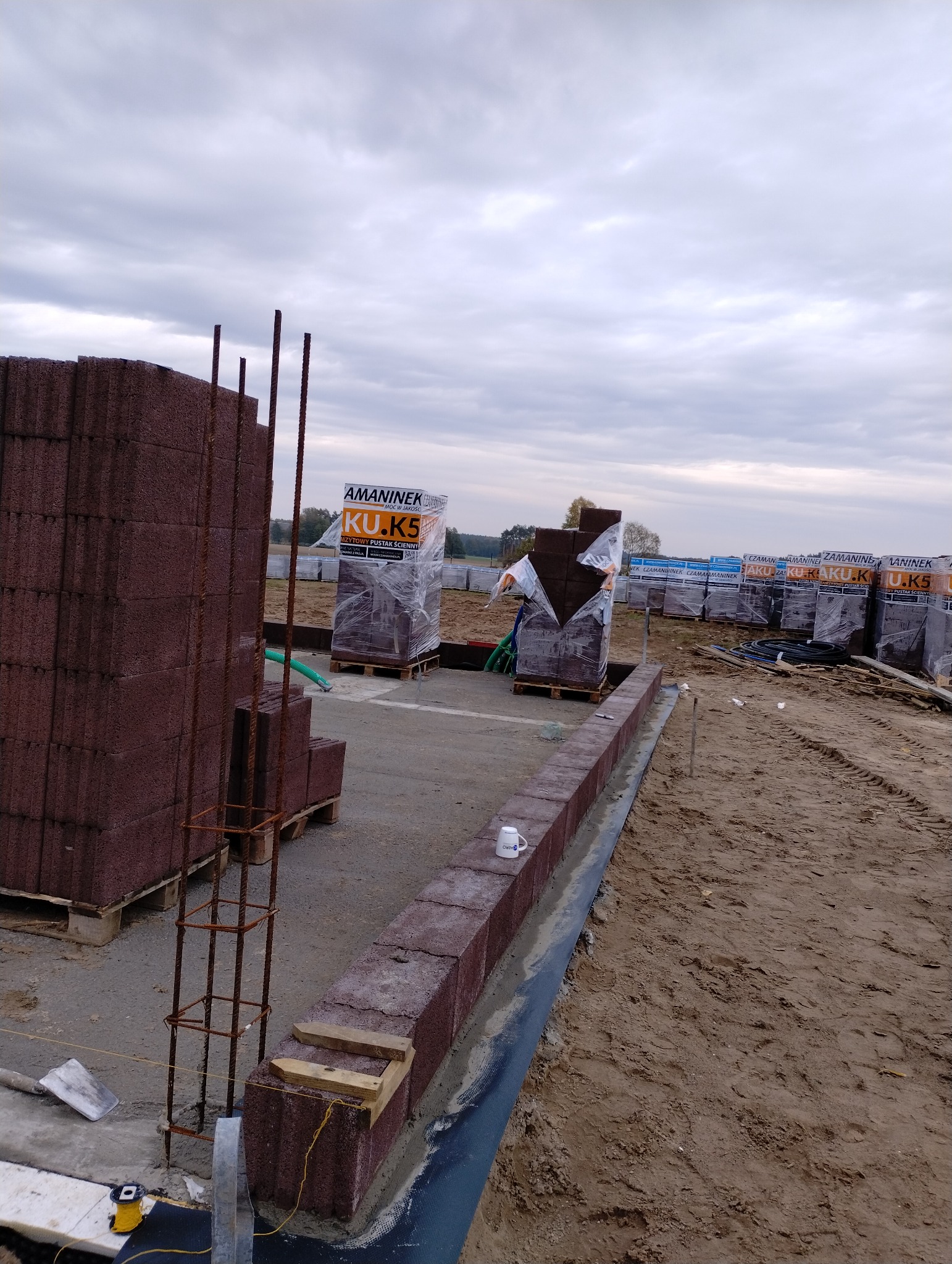 Budowa ogrodzenia z brązowych bloczków betonowych, widoczne zbrojenie narożnika, palety z zapakowanymi bloczkami w tle, kubek na murku.