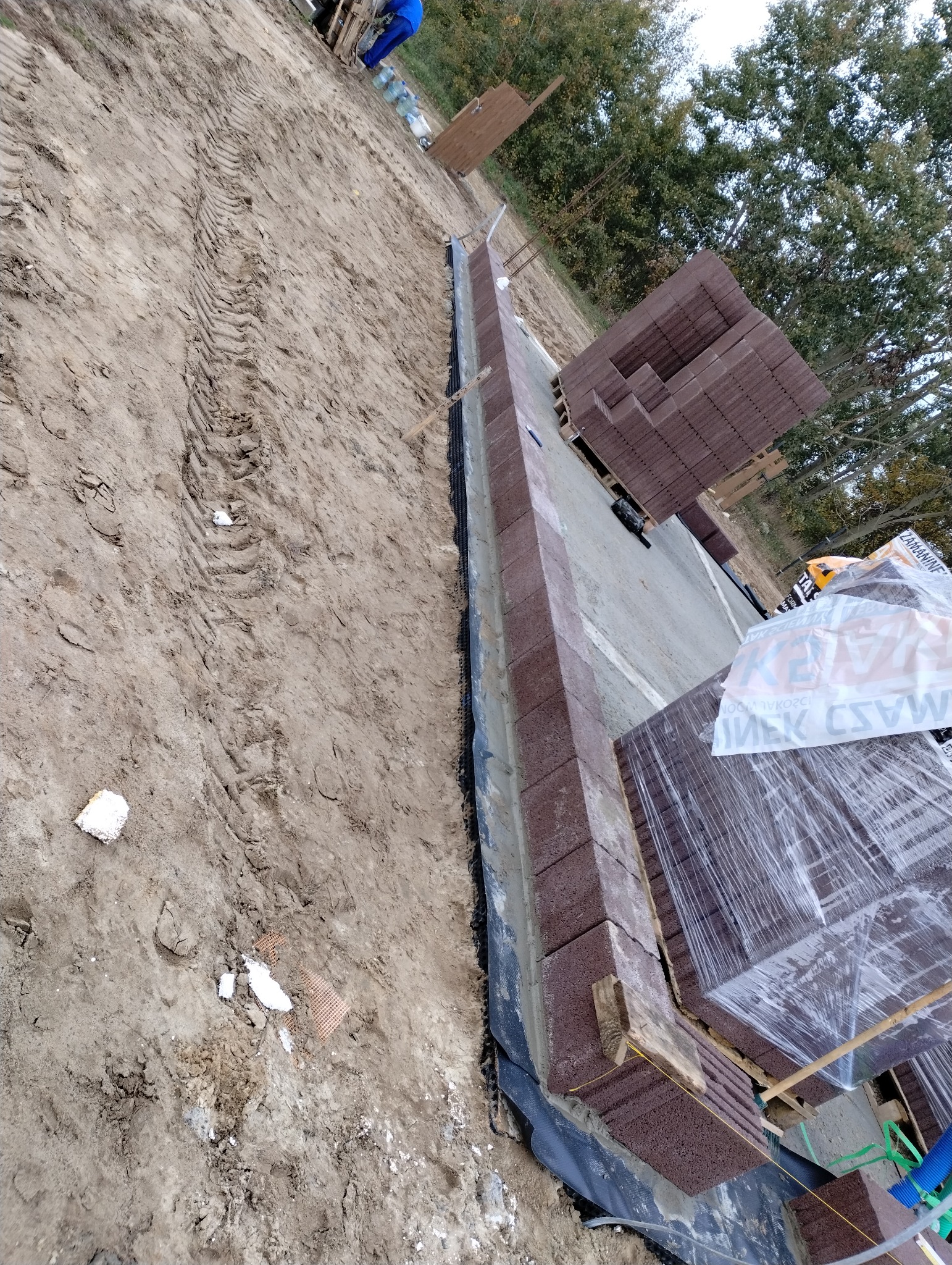 Wykop pod fundament obłożony kostką brukową w kolorze ceglastym, zabezpieczony czarną folią. Na palecie obok ułożone kostki brukowe w folii. Widoczne ślady opon w ziemi.