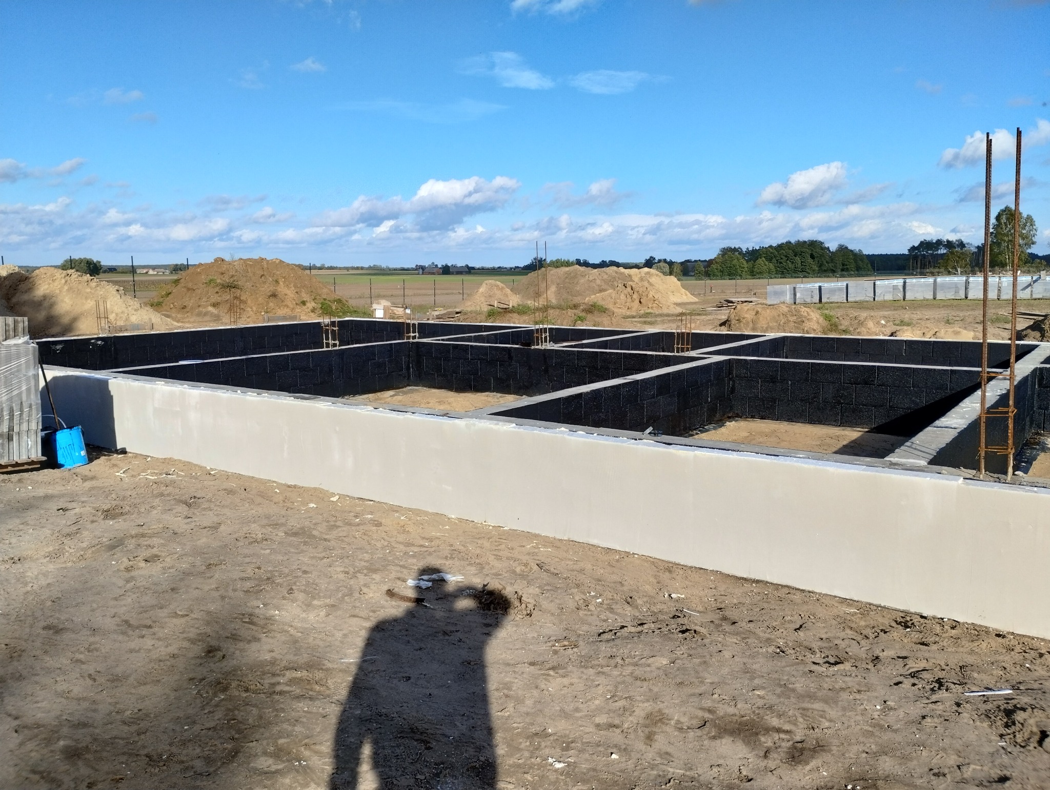 Fundamenty domu jednorodzinnego z bloczków betonowych, z zewnątrz otynkowane na gładko, z widocznymi prętami zbrojeniowymi, na tle piaszczystego terenu i błękitnego nieba z chmurami. W tle hałdy...