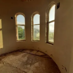 Wnętrze okrągłego pomieszczenia z trzema łukowatymi oknami w trakcie tynkowania maszynowego, widoczne niedoskonałości i rury na podłodze.