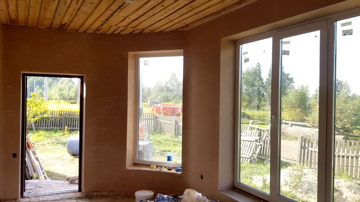 Wnętrze domu w trakcie tynkowania maszynowego: surowy tynk na ścianach, drewniany strop, białe okna z widokiem na zewnątrz, widoczne niedoskonałości i ślady prac budowlanych.