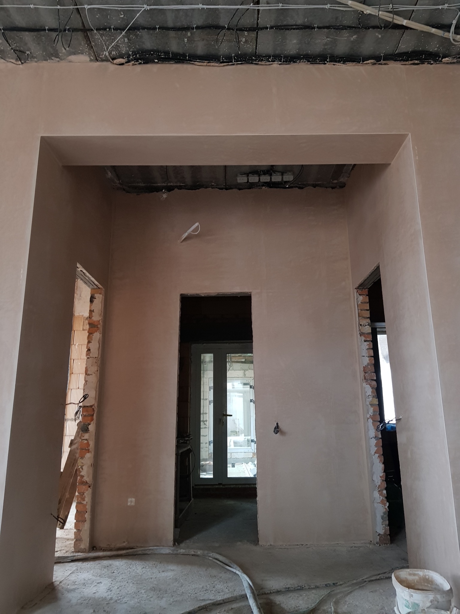 Świeżo nałożony, gładki tynk na ścianach wewnętrznych budynku w trakcie remontu, widoczne przewody elektryczne pod sufitem i surowe ościeżnice drzwiowe.