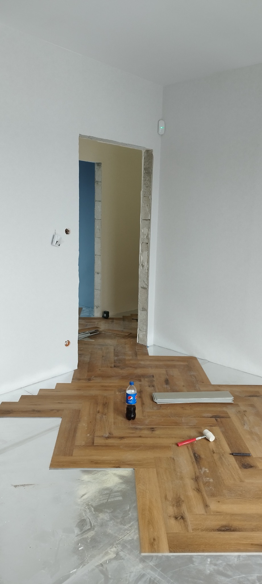 Układanie paneli podłogowych w jodełkę w pomieszczeniu w trakcie remontu, widoczna butelka napoju, młotek gumowy i listwy wykończeniowe.