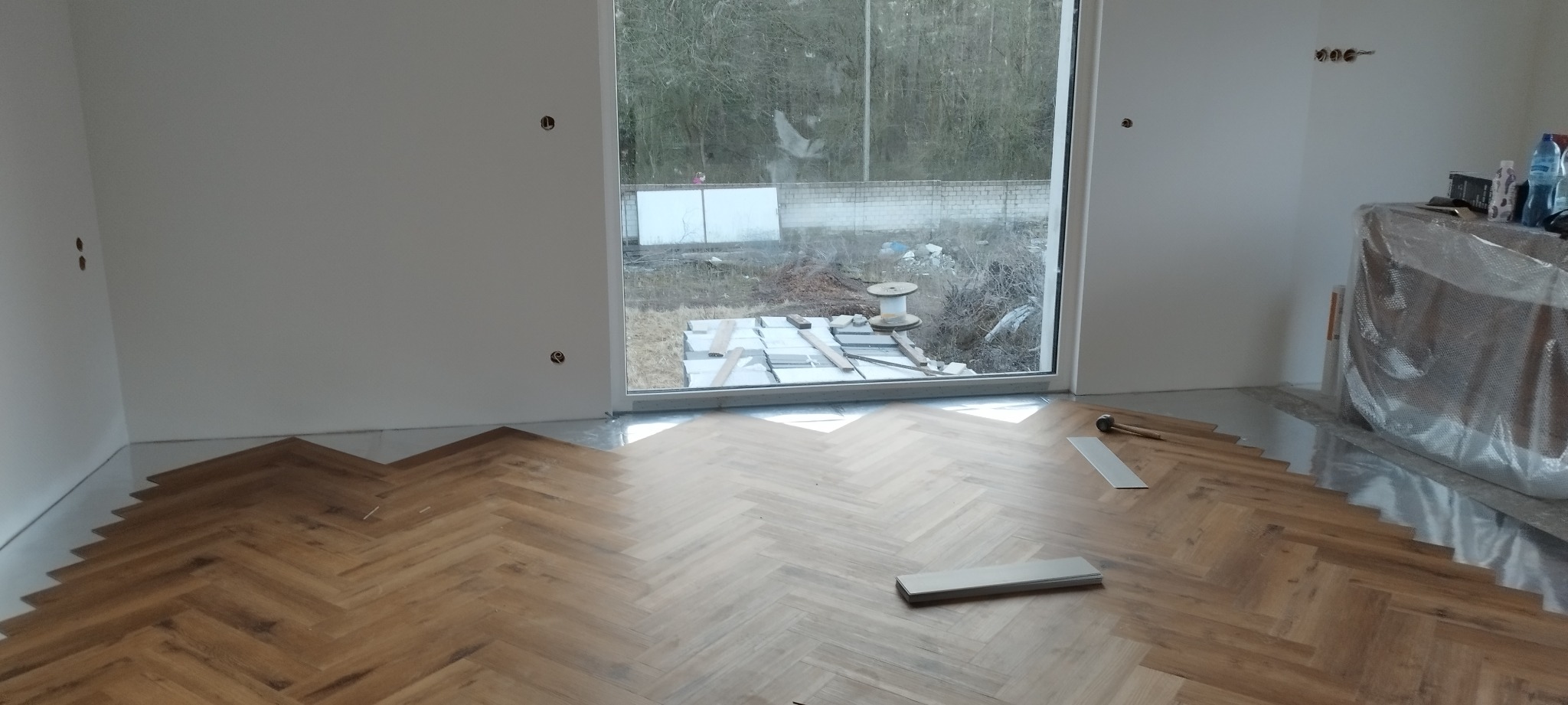 Fragment układania parkietu w jodełkę w jasnym pomieszczeniu z dużym oknem, widoczne niedokończone krawędzie i narzędzia pracy.