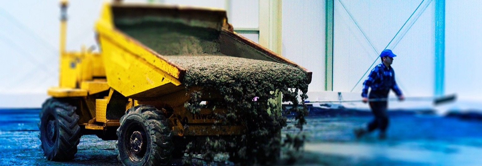 Żółta wywrotka z betonem wylewająca zawartość na niebieską posadzkę. W tle pracownik z miotłą. Ujęcie z bliska na koła i wylewający się beton.