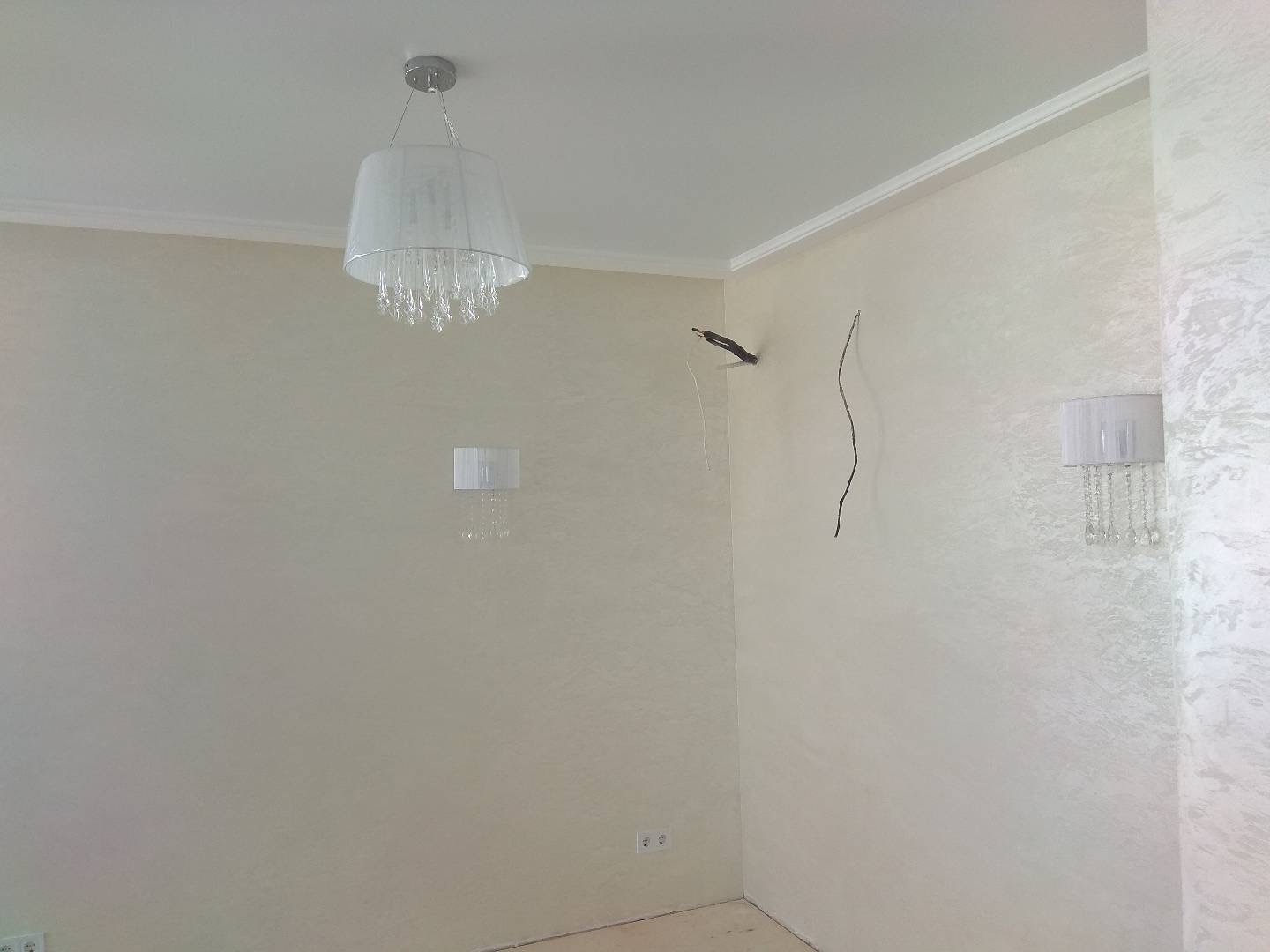 Wnętrze pokoju z zamontowanym żyrandolem sufitowym i kinkietem ściennym, widoczne przewody elektryczne wystające ze ściany, ściany pokryte dekoracyjnym tynkiem.