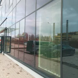 Szklana fasada biurowca w Jeleniej Górze z odbiciem samochodów i przemysłowego krajobrazu. Widoczne regularne podziały okien i chodnik z kostki brukowej.