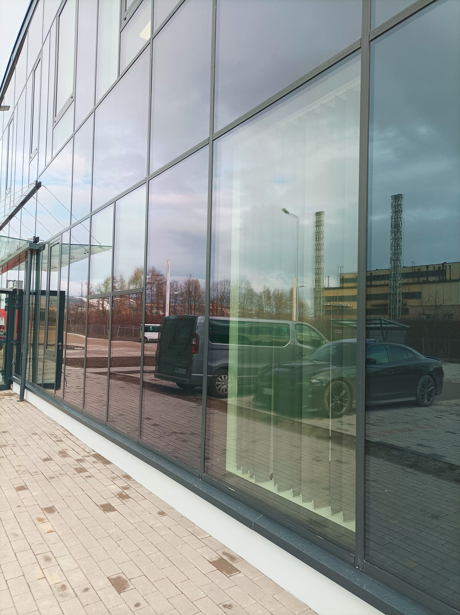 Szklana fasada biurowca w Jeleniej Górze z odbiciem samochodów i przemysłowego krajobrazu. Widoczne regularne podziały okien i chodnik z kostki brukowej.