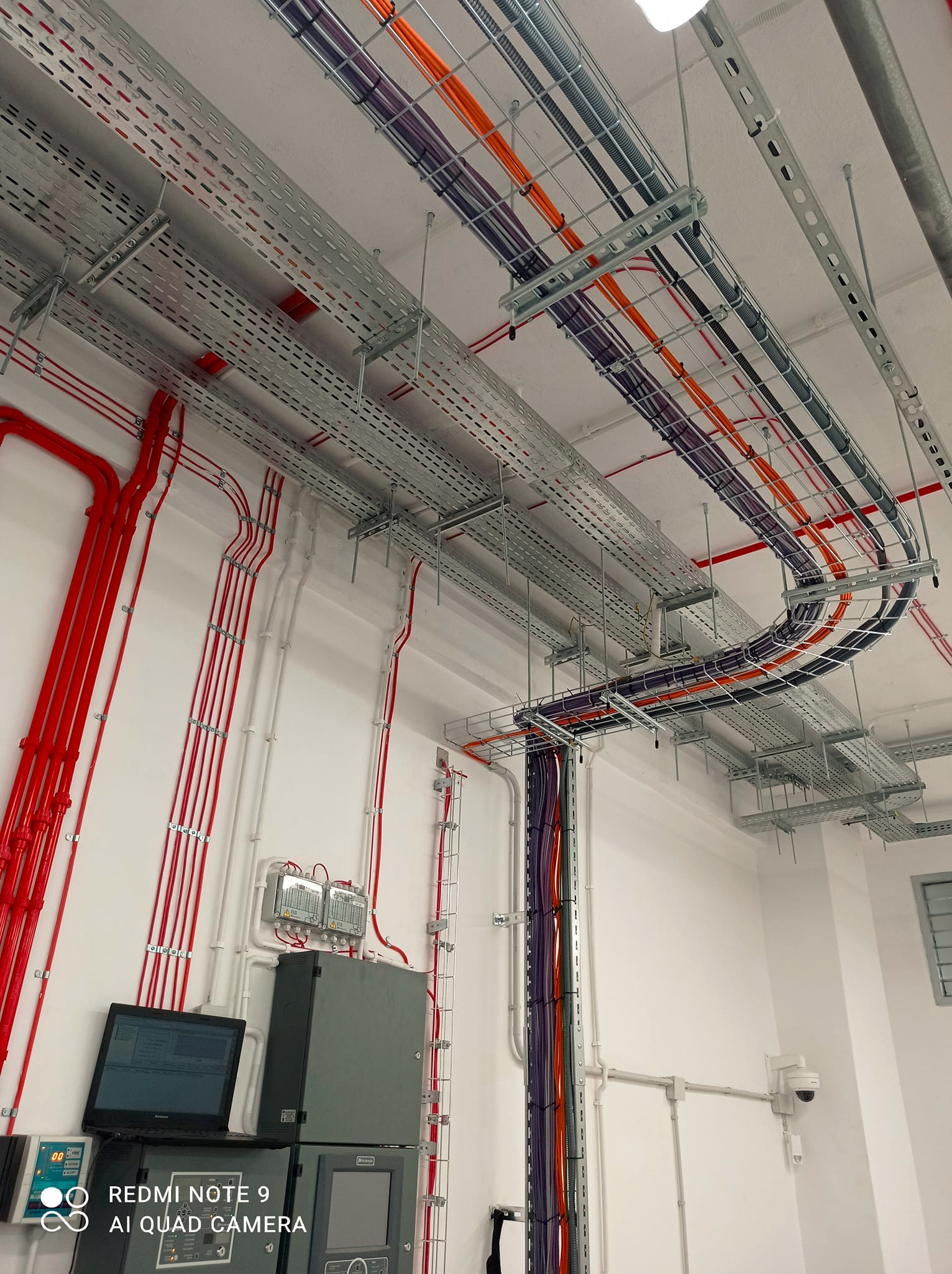 Instalacja elektryczna i teletechniczna w budynku: koryta kablowe z przewodami w kolorach pomarańczowym, fioletowym i czarnym pod sufitem, pionowe rury z czerwonymi kablami na ścianie, szafa...