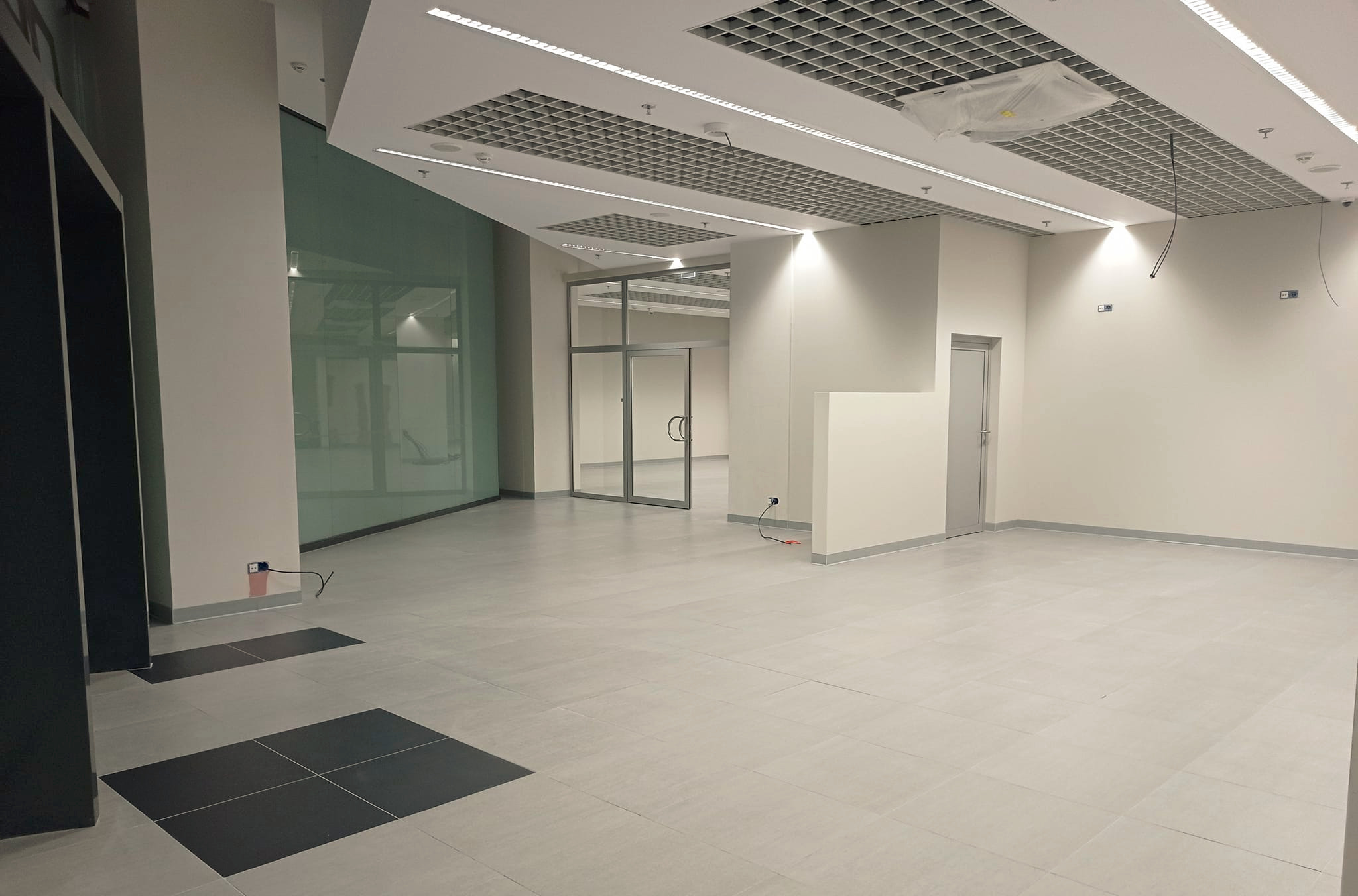 Puste, jasne pomieszczenie biurowe z szarą podłogą z płytek, przeszkloną ścianą i podwieszanym sufitem z kratką, gotowe do zagospodarowania.