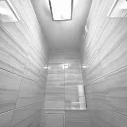 Wąska toaleta w odcieniach szarości z płytkami imitującymi marmur, oświetleniem sufitowym i rolkami papieru toaletowego na półce.