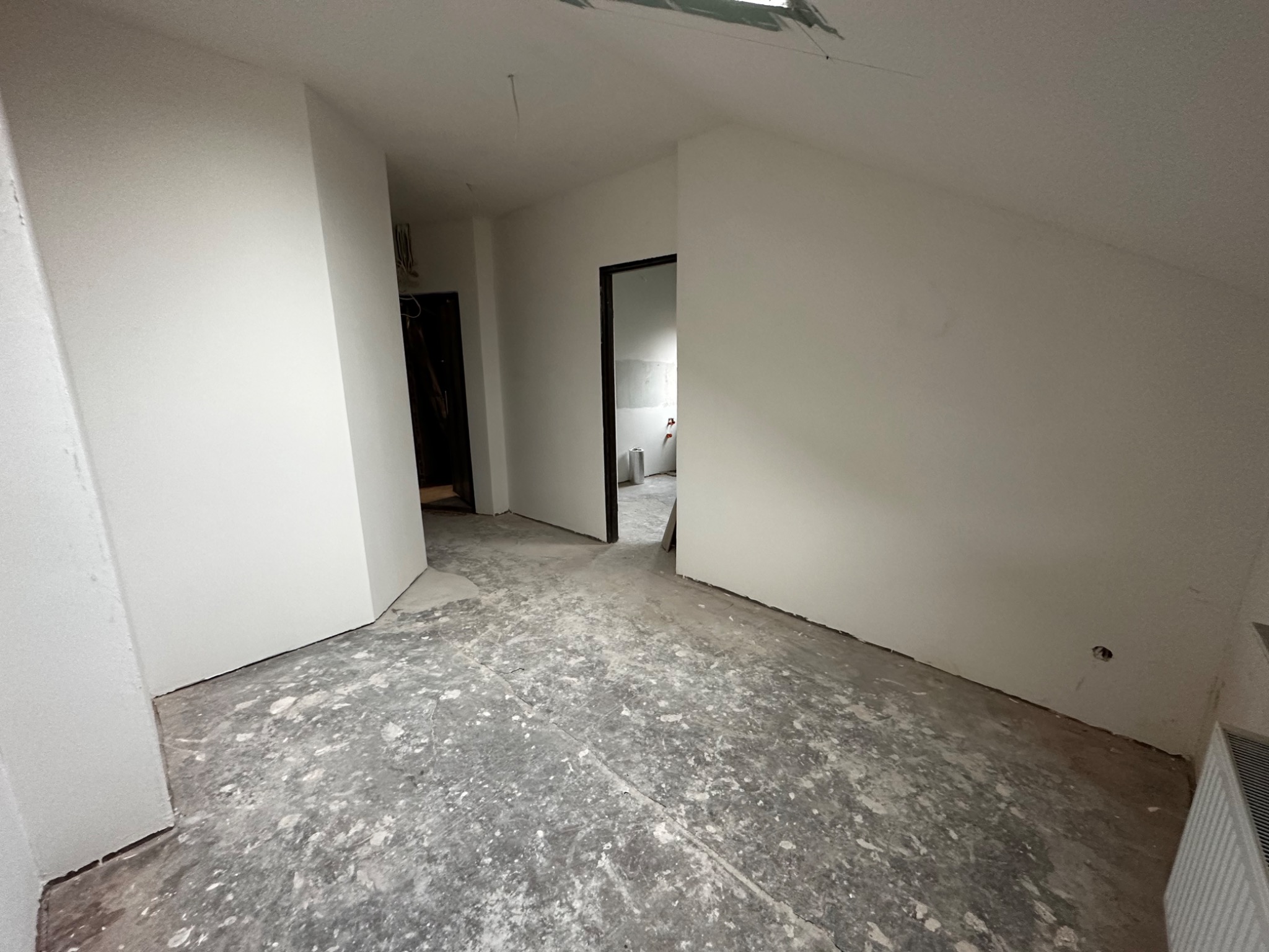 Poddasze w trakcie prac wykończeniowych; świeżo pomalowane ściany na biało, surowa podłoga z widocznymi zabrudzeniami po farbie, widoczne otwory drzwiowe.