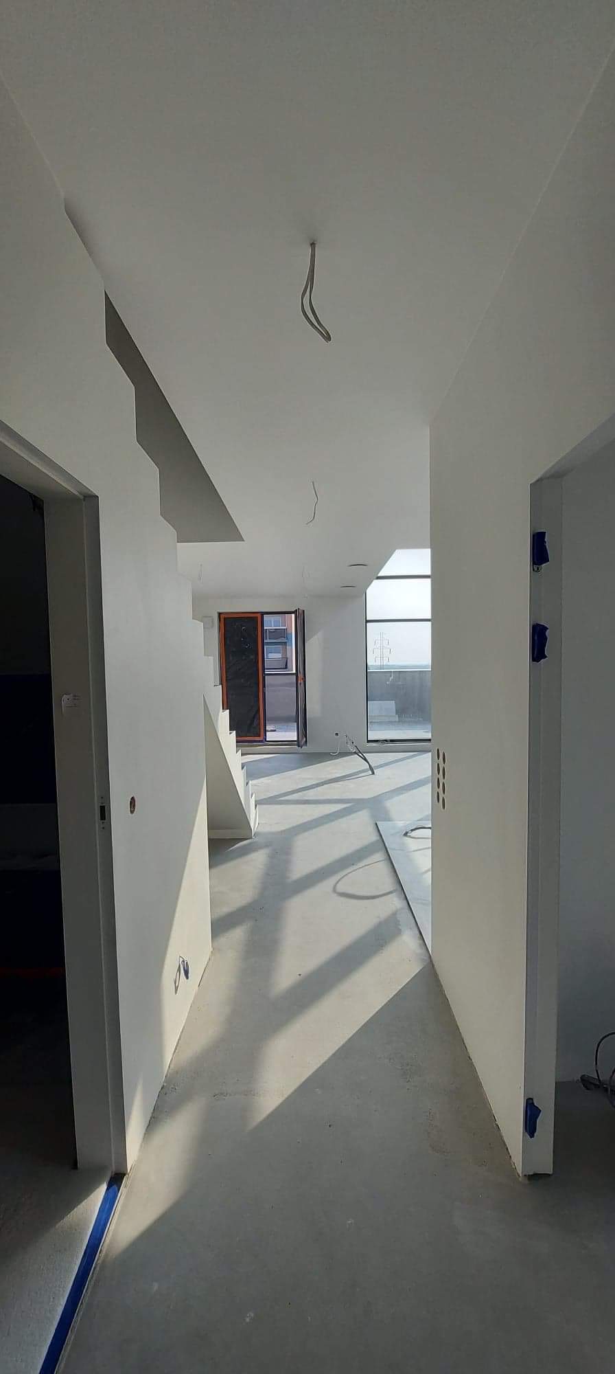 Wnętrze w trakcie prac wykończeniowych: surowe ściany pomalowane na biało, widoczne przewody wystające z sufitu, zabezpieczona taśmą podłoga i fragment schodów w jasnym, słonecznym pomieszczeniu.