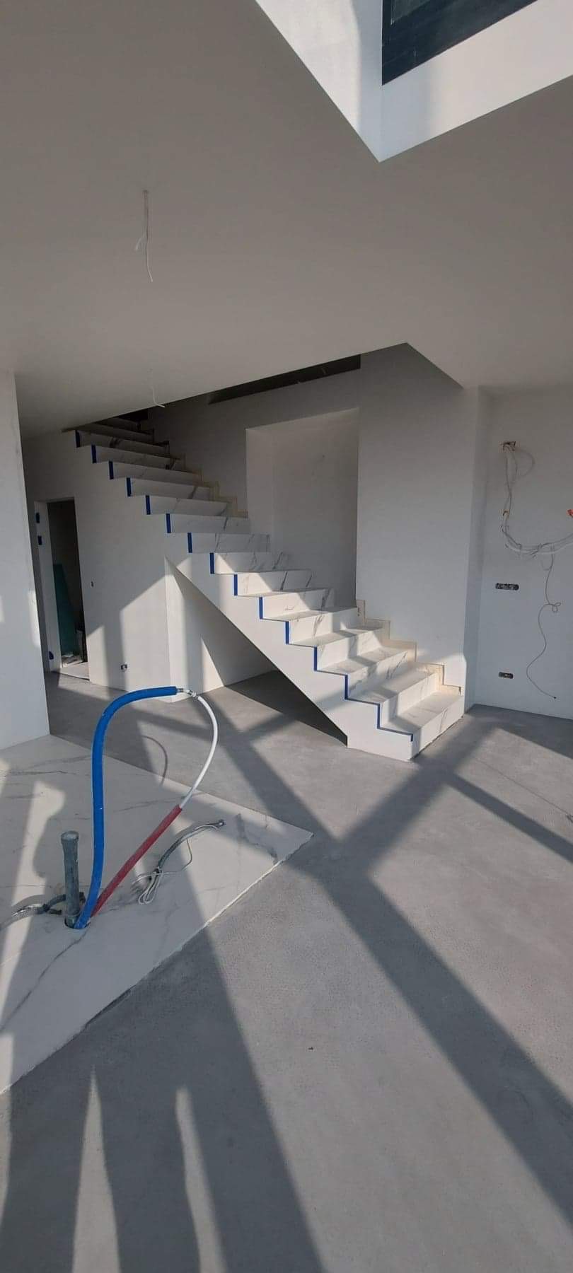 Nowoczesne, białe schody z marmurowymi stopniami, zabezpieczone niebieską taśmą malarską, w surowym wnętrzu z widocznymi instalacjami hydraulicznymi i elektrycznymi, w trakcie wykańczania.
