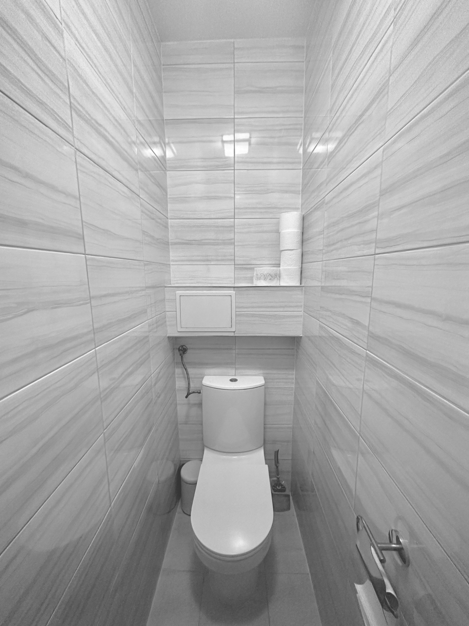 Wąska toaleta wykończona jasnymi, marmurkowymi płytkami, z podwieszaną muszlą klozetową i ukrytym zbiornikiem, na półce zapas rolek papieru toaletowego.