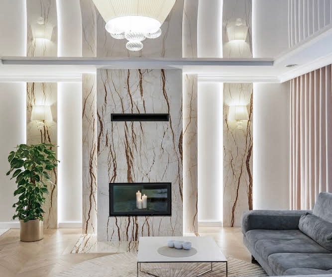 Elegancki salon z marmurowym kominkiem, nowoczesnym oświetleniem sufitowym i ścianami z podświetleniem LED, uzupełniony szarą sofą i rośliną doniczkową.