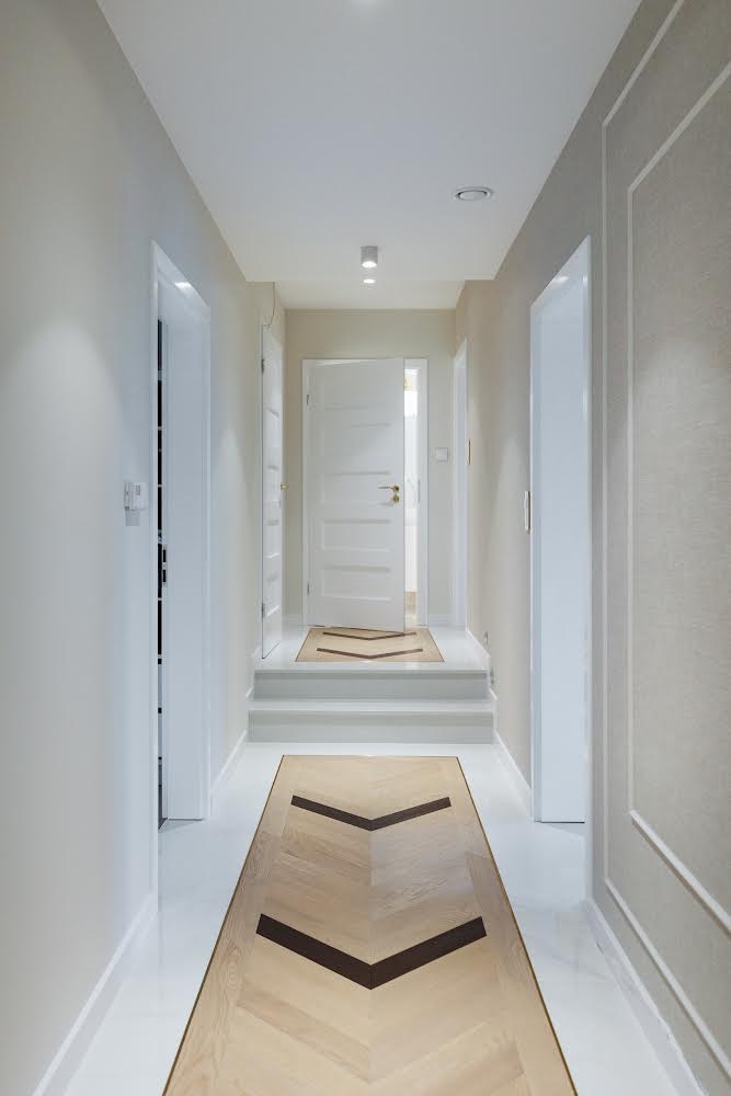 Nowoczesny korytarz w Radomiu z jasnymi ścianami, białymi drzwiami i podłogą z wzorem w jodełkę, oświetlony punktowymi lampami sufitowymi.