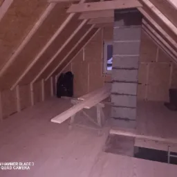 Poddasze z oknem w trakcie zabudowy płytami OSB, widoczny komin z bloczków betonowych, stół roboczy z deskami i czarny worek na śmieci.