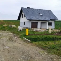 Nowo wybudowany, dwukondygnacyjny dom murowany z ciemnoszarą dachówką, brązową bramą garażową i żółtymi słupkami gazowymi na nierównym terenie.