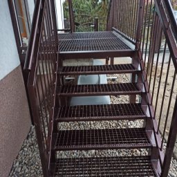 LAKRYS Krystian Lalak - Zewnętrzne metalowe schody z kratką pomalowane na brązowo, prowadzące do góry, otoczone drobnymi kamieniami.