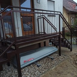 LAKRYS Krystian Lalak - Zewnętrzne metalowe schody z podestem prowadzące do drzwi, z szarym boxem dachowym pod spodem, ułożonym na żwirze.