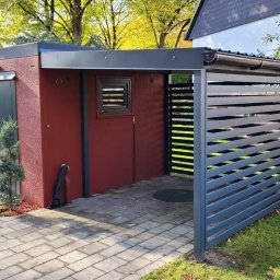 LAKRYS Krystian Lalak - Nowoczesna wiata garażowa z elementami zadaszenia i ażurowej przesłony w odcieniach szarości i bordo, z kostki brukowej ułożonej na podjeździe, z psem rasy mieszanej obserwującym otoczenie.